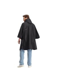 Zwarte waterdichte poncho met een capuchon, met een losse pasvorm, zijsplitten en reflecterende accenten. Gecombineerd met lichtblauwe jeans en beige schoenen.