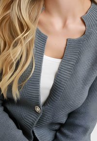 Donna bionda che indossa un cardigan grigio a trama con un bottone argentato su una maglietta bianca, mostrando il collo e i lunghi capelli mossi.