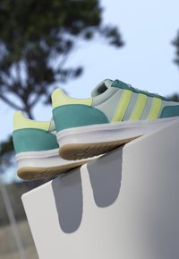 Paar grüne und gelbe Sneaker mit weißen Sohlen, die am Rand einer weißen Fläche vor einem verschwommenen Außenhintergrund platziert sind.