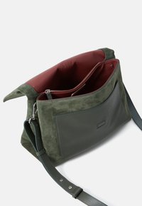Borsa a tracolla in suede verde e pelle con una tasca con zip e interni di un rosso profondo. Presenta una tracolla regolabile e una tasca esterna.