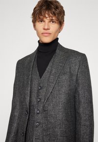 Blazer gris texturizado con solapa muesca y dos botones, superpuesto a un cuello alto negro. Presenta un patrón sutil y botones en contraste.