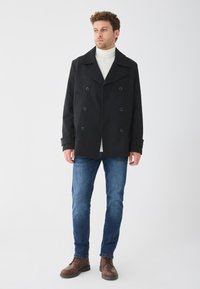 Doppelreihiger schwarzer Mantel mit breiten Rever, strukturiertem Stoff, seitlichen Taschen und großen Knöpfen, kombiniert mit blauen Jeans und braunen Stiefeln.