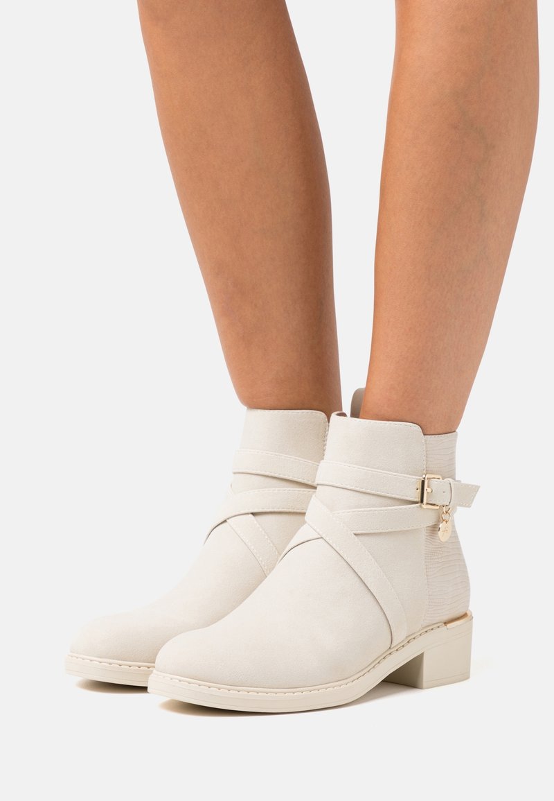 Bottes crème montant à la cheville avec des sangles croisées, une boucle latérale ornée d'un petit charme, et un talon bloc bas portées sur des jambes nues.