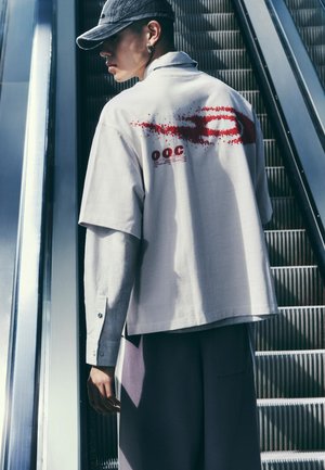 Persoon met een grijze pet en gelaagde witte en gestreepte shirts met een rode print op de rug, staand naast een metalen roltrap.