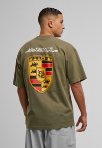 Oliven grøn oversized t-shirt med et stort gult og sort skjoldlogo på ryggen samt fed tekst ovenfor. Korte ærmer, lavet af bomuldsmateriale.