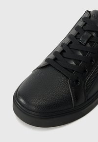 Baskets en cuir noir texturé avec lacets noirs et détail de fermeture éclair latérale sur fond gris clair.