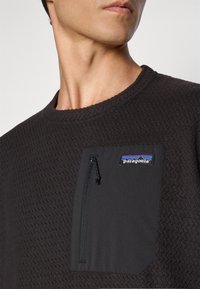 Gros plan sur une personne portant une veste polaire noire texturée de Patagonia avec une poche poitrine zippée et un patch de logo.