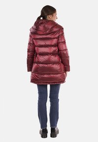Parka di piuma color bordeaux, con cappuccio, texture trapuntata e un taglio leggermente più lungo. Abbinata a jeans blu e calzature nere.