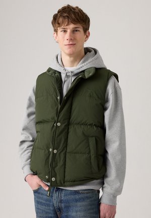 Zielona kamizelka puchowa z zatrzaskami, przednimi kieszeniami i wysokim kołnierzem, noszona na szarym hoodie. Materiał wydaje się gładki i lekko teksturowany.