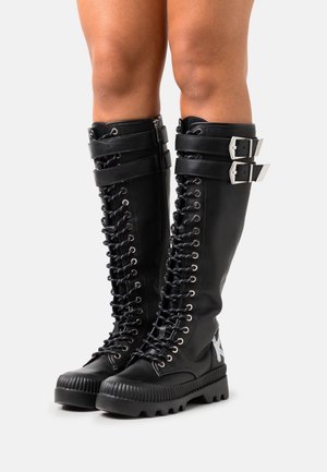 Zwart leren kniehoge laarzen met een voorzijde met veters, zilverkleurige hardware, twee verstelbare riemen en een gestructureerde zool.