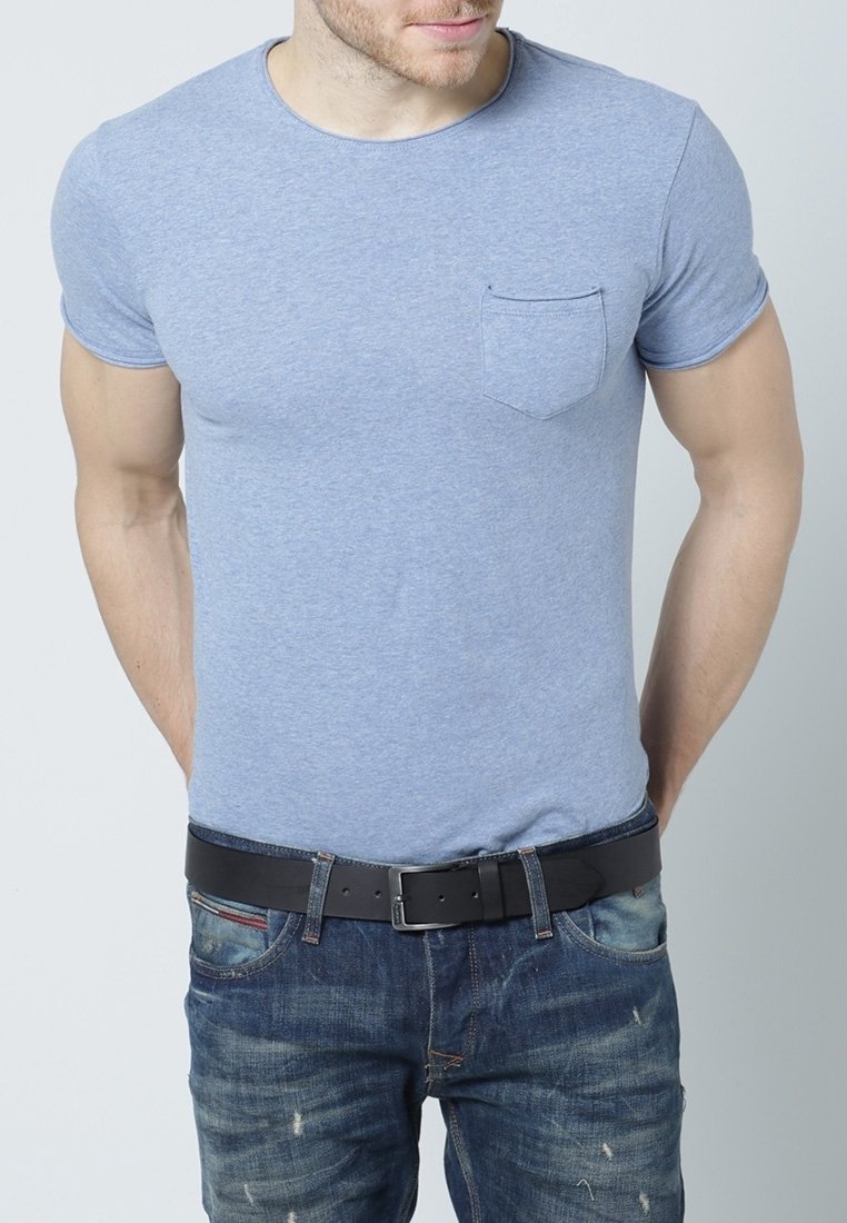 Homme portant un t-shirt ajusté bleu clair à manches courtes avec poche poitrine et un jean bleu usé avec ceinture en cuir noire, mains dans le dos.