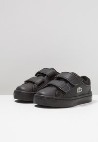 Černé kožené boty se dvěma nastavitelnými popruhy na suchý zip, kulatou špičkou, silnou černou gumovou podrážkou a malým zeleným vyšitým logem krokodýla.