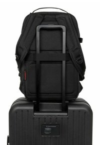 Svart ryggsäck med diamantmönstrad tyg vilar på en svart hårdskalväska med vertikala spår och en metallidentifieringsskylt.