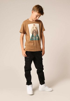 Bruine t-shirt met een hondenprint, gecombineerd met zwarte cargobroek met zakken. Witte sneakers maken de outfit compleet. Eenvoudige achtergrond.