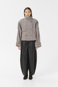 Kvinne iført høyhalset houndstooth jakke med knappdetaljer og brede svarte skinnbukser, som står foran en hvit bakgrunn.