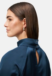 Navy Satin-Bluse mit hohem Kragen, Rückenausschnitt und zwei Knopfakzenten. Der Stoff hat eine glatte, glänzende Textur.