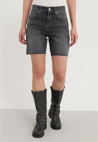 Shorts de mezclilla negra con dobladillo sin acabar, bolsillos frontales y una cintura alta, combinados con botas de cuero negras hasta la rodilla con detalles de hebilla.