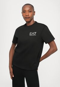 Person med kort hår iført en svart kortermet t-skjorte med hvit "EA7 Emporio Armani"-logo, som står med en hånd bak ryggen.