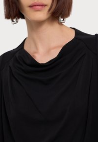 Schwarzes Oberteil mit drapiertem Ausschnitt und weicher Textur, das dezente Fältchen und eine lockere Passform aufweist. Sichtbares silbernes Hardware-Detail.