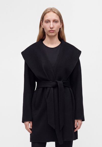 Femme aux longs cheveux blonds portant un manteau noir ceinturé avec de larges revers, debout devant un fond clair uni.