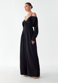 Moteris vilki juodą suknelę be petnešų, plačiomis kelnėmis, su auksinėmis auskarų kilpelėmis ir atvirais aukštakulniais bateliais, stovi prieš paprastą baltą foną.