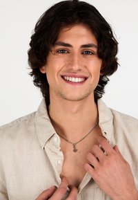 Collana con una catena d'argento che presenta un piccolo pendente a forma di ancora; il modello indossa una camicia con colletto color beige chiaro, mettendo in evidenza chiaramente i gioielli.