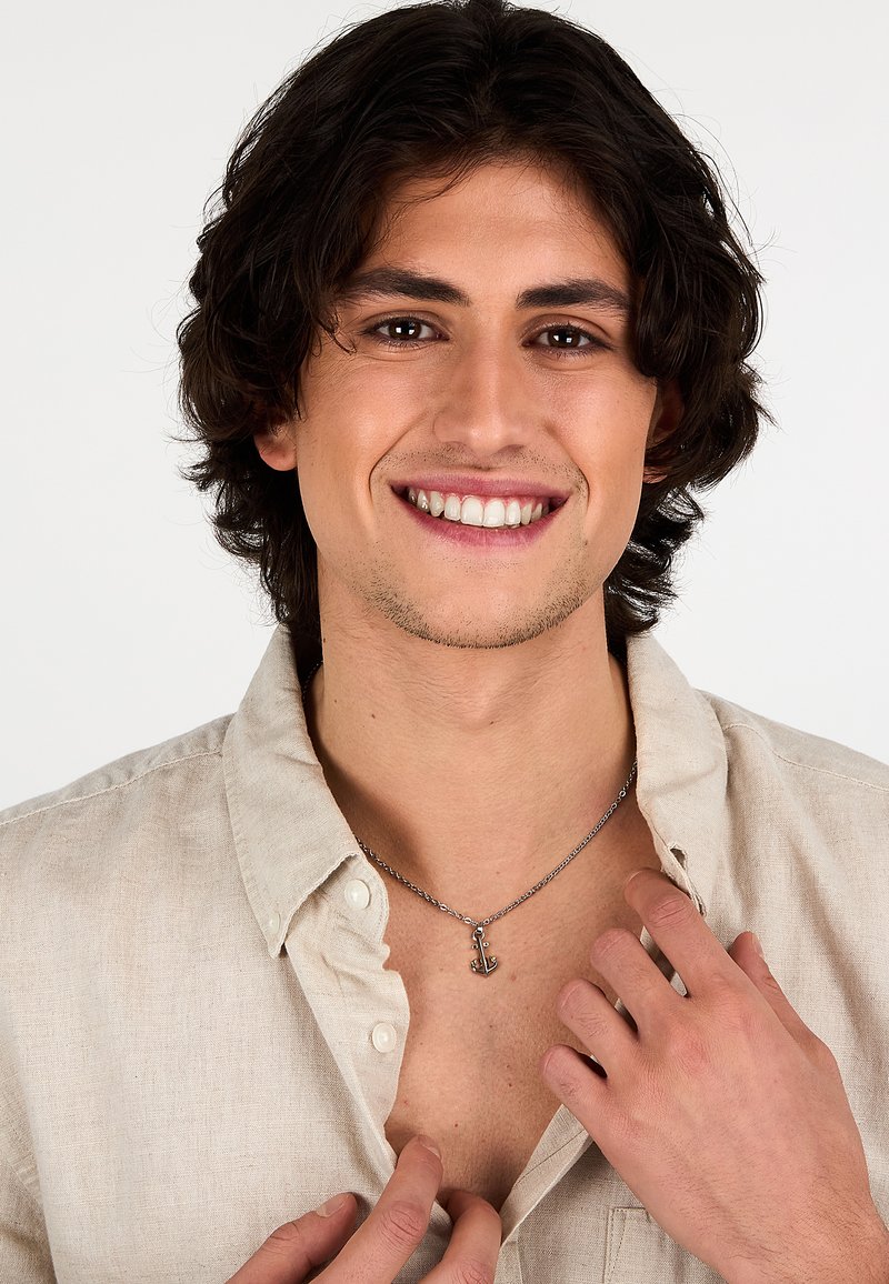Collana con una catena d'argento che presenta un piccolo pendente a forma di ancora; il modello indossa una camicia con colletto color beige chiaro, mettendo in evidenza chiaramente i gioielli.