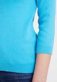 Ljusa blå långärmad tröja med ribbade muddar och kant bärs över mörkblå jeans på en persons arm och torso.