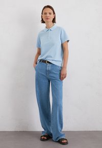 Lichtblauwe poloshirt met korte mouwen, gecombineerd met brede spijkerbroek van denim en zwarte sandalen, tegen een neutrale achtergrond.