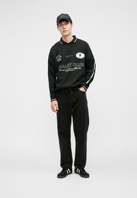 Polo a maniche lunghe nera con dettagli bianchi e grafiche ricamate, abbinato a jeans scuri e sneakers nere. Sfondo semplice.