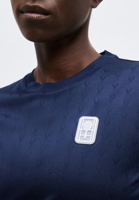 Κοντινό πλάνο ενός ατόμου που φοράει μπλε σκούρο πουκάμισο με διακριτικό υφή μοτίβο και λευκό έμβλημα ρακέτας τένις στο στήθος.