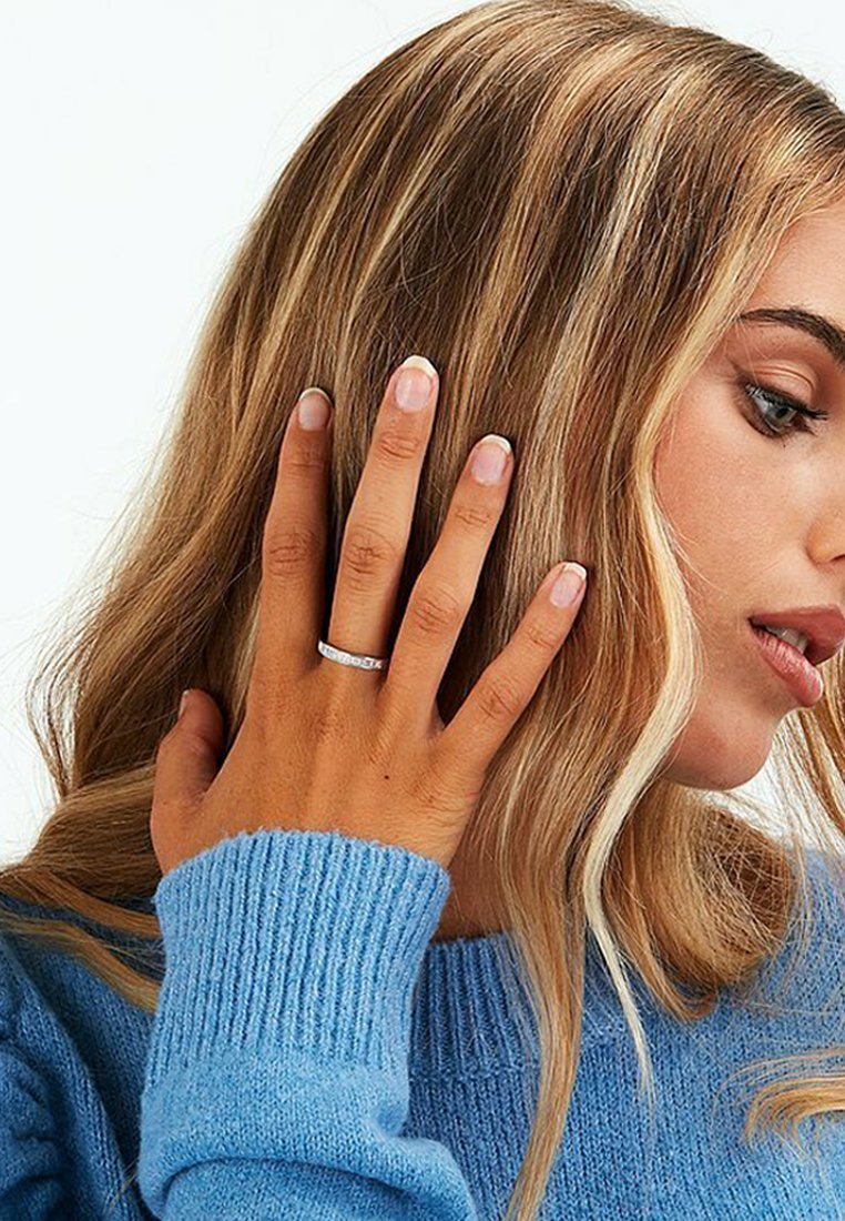 Silberner Ring mit einem schlichten Band und kleinen klaren Steinen, getragen an einer Hand neben sanftem, welligem Haar, das einen blauen gerippten Pullover präsentiert.