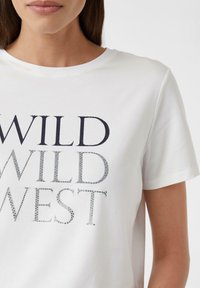 Weißes Kurzarm-T-Shirt mit dem Aufdruck "WILD WILD WEST" in fetten Buchstaben, verziert mit silbernen Nieten für zusätzliche Textur und Details.