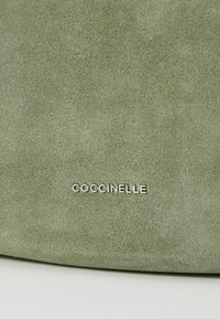 Green suede bag with a smooth texture and a silver "Coccinelle" logo prominently displayed. The design is simple and elegant.