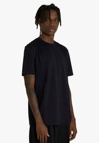 Giovane uomo con dreadlocks che indossa una maglietta nera semplice e pantaloni neri, in piedi con un'espressione neutra contro uno sfondo bianco.