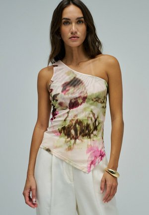 Femme portant un top fleuri asymétrique à une épaule et un pantalon blanc taille haute, accessoirisée d'un large bracelet en or.