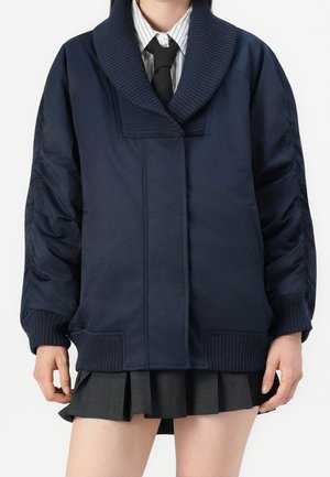 Personne portant une veste bleu marine oversize avec col et poignets côtelés, une chemise rayée, une cravate noire et une jupe plissée foncée.