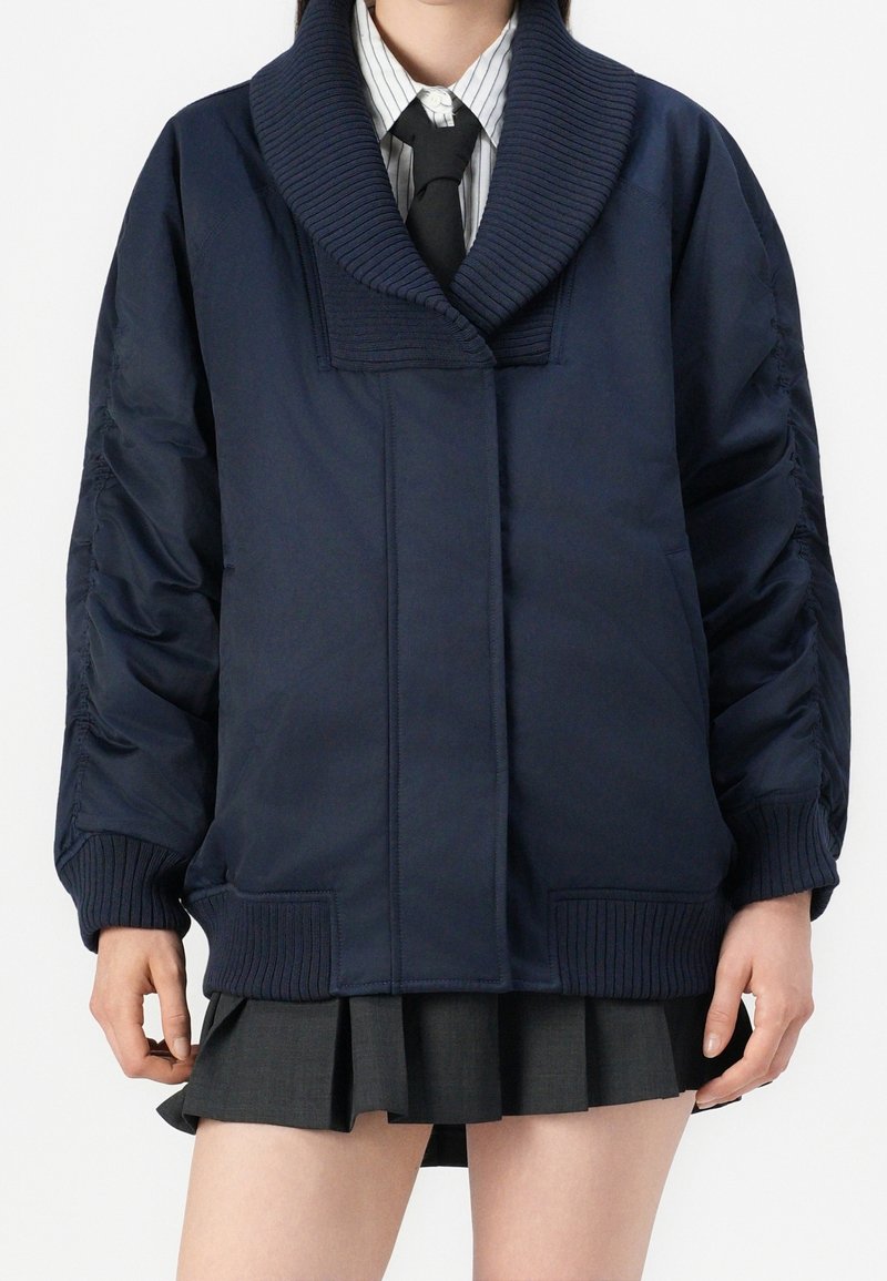 Personne portant une veste bleu marine oversize avec col et poignets côtelés, une chemise rayée, une cravate noire et une jupe plissée foncée.