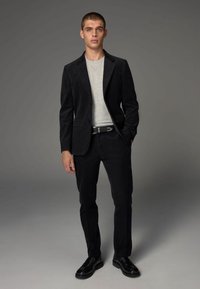 Schwarzer Cordanzug mit tailliertem Schnitt, kombiniert mit einem grauen T-Shirt. Schwarze Schuhe und ein eleganter Gürtel runden das Outfit ab.