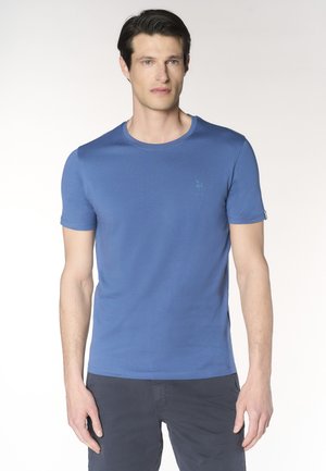 Uomo con capelli scuri che indossa una maglietta blu tinta unita a maniche corte e pantaloni scuri, in piedi davanti a uno sfondo chiaro.