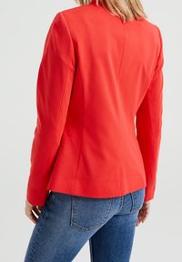 Red tailored blazer with long sleeves, structured shoulders, and a fitted silhouette, paired with blue denim jeans. Smooth fabric texture.