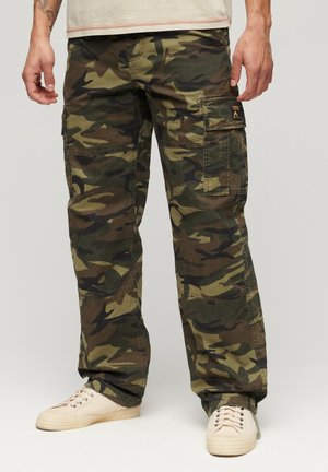 Mann trägt grüne und braune Tarn- Cargo-Hose mit beigen Sneakers vor einem einfarbigen grauen Hintergrund.