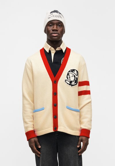 Κρεμ ζακέτα με κόκκινες λεπτομέρειες, μπλε καλύμματα τσέπης, μαύρα κουμπιά και κέντημα αστροναύτη αριστερά στο στήθος· φοριέται με μαύρο πουκάμισο και μπεζ σκούφο.