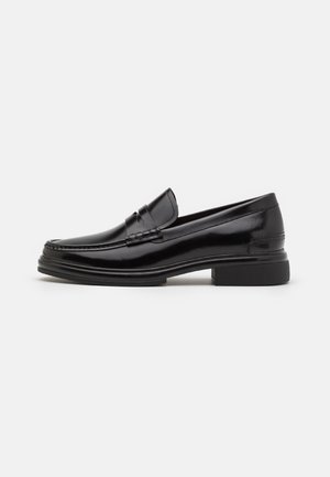 Mocassins en cuir noir avec un embout arrondi, présentant un détail cousu sur le dessus, et un petit talon carré. Texture lisse et finition brillante.