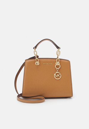 Beżowa torebka Michael Kors z złotym łańcuszkowym uchwytem, zawieszką z logo MK oraz cienkim, regulowanym paskiem na ramię na białym tle.