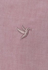 Tela texturizada rosa con un colibrí bordado en hilo gris claro, con un diseño detallado en las alas y el cuerpo.
