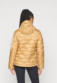 Dikke tan kleurige jas met een getextureerd golfpatroon, een vaste capuchon, elastische manchetten en een lichte accentkleur op de linkerschouder.