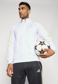 Hvid sportjakke med lyseblå og lyserøde detaljer, gule striber på ærmerne, parret med mørke bukser. Holder en sort-hvid fodbold.