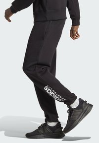 Pantaloni sportivi neri con dettagli del logo lungo la gamba sinistra, abbinati a scarpe sportive nere e calze bianche, realizzati in un tessuto testurizzato.