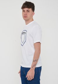 T-shirt bianca a maniche corte con un design a scudo blu sul davanti. Realizzata in cotone, presenta un colletto classico e una vestibilità relax.